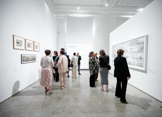 Baltā muzeja izstāžu zālē apmeklētāji apskata pie sienām izvietotus grafikas un zīmējumus rāmjos.