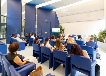 Cilvēki sēž modernā konferenču telpā ar zilām sienām un klausās prezentāciju, kas tiek rādīta uz ekrāna.