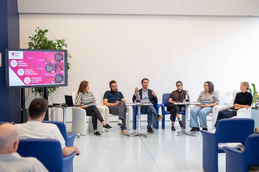 A discussion panel with six speakers seated in a semi-circle in a conference room, next to a screen showing a presentation on cultural heritage and technology.