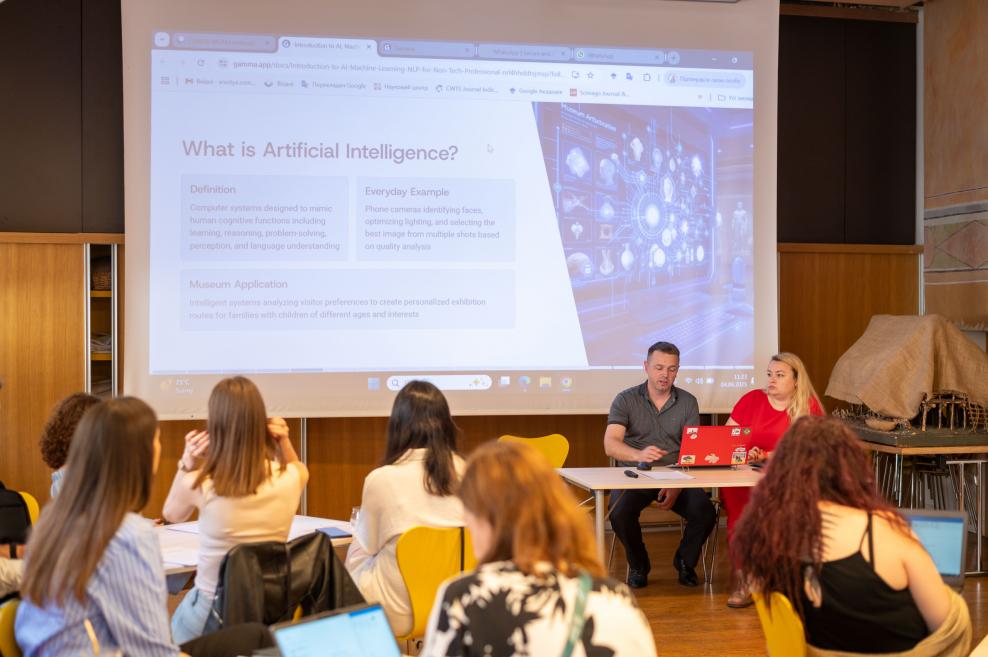 Lekcija par mākslīgo intelektu notiek klasē, kur priekšā sēž lektori ar portatīvajiem datoriem un uz ekrāna redzama prezentācija ar tēmu ‘What is Artificial Intelligence?’
