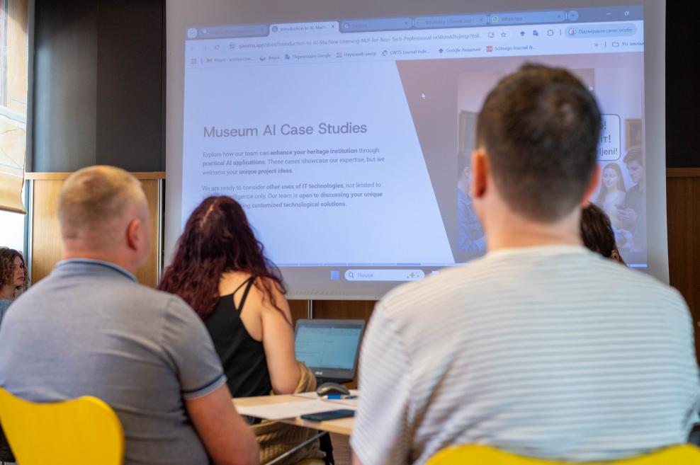 Dalībnieki klausās prezentāciju par mākslīgā intelekta pielietojumu muzejos, skatoties uz projekciju ar virsrakstu ‘Museum AI Case Studies’.