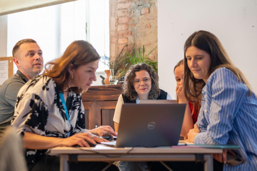 Grupa dalībnieku sēž ap galdu un strādā pie datora neformālā semināra vidē ar ķieģeļu sienu fonā.