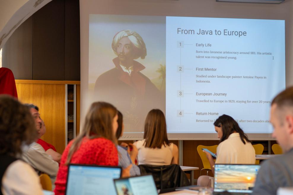 Dalībnieki piedalās lekcijā, kurā uz ekrāna redzama prezentācija par mākslinieka ceļojumu ‘From Java to Europe’ un viņa biogrāfija.