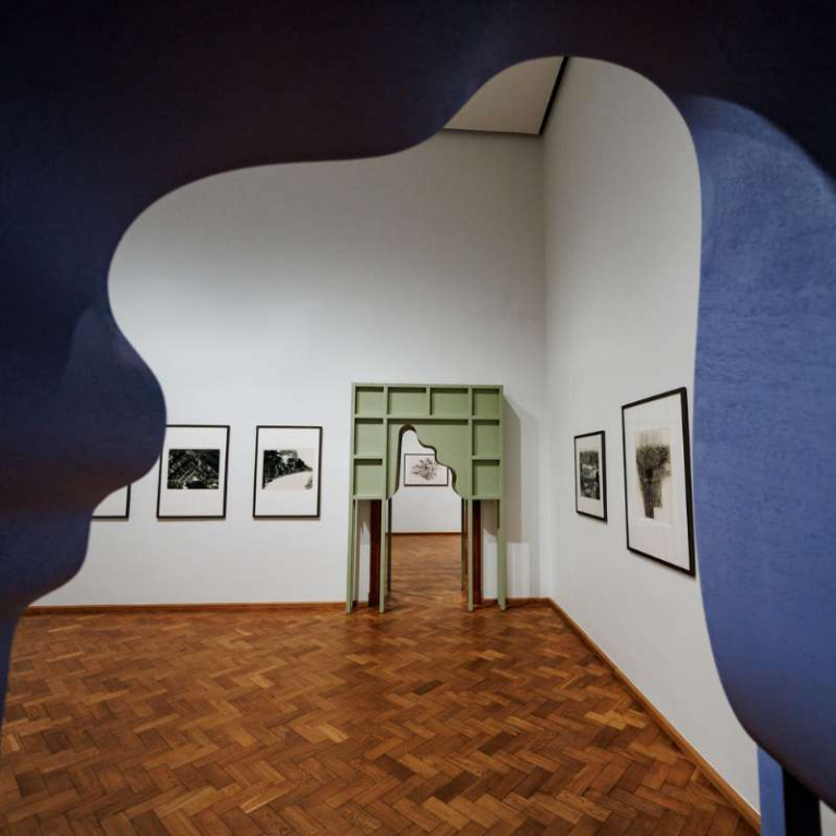 View down a museum corridor with parquet flooring and columns, seen through the opening of a large white sculpture that frames the perspective toward the far end of the hall.