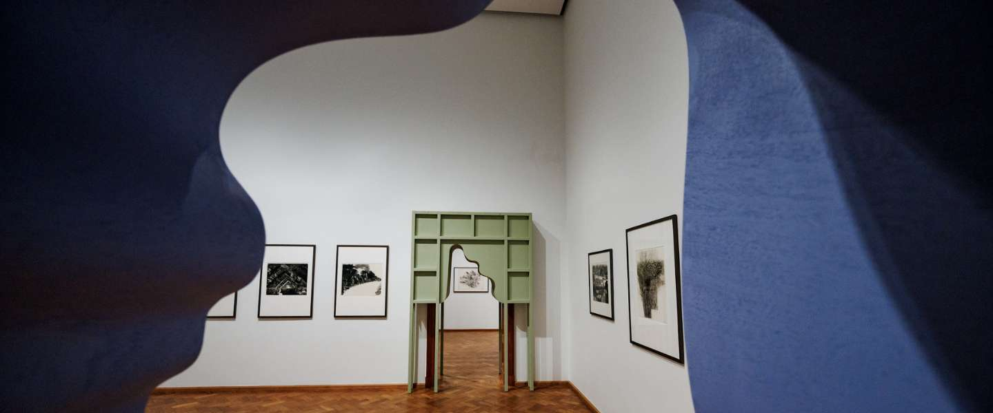 View down a museum corridor with parquet flooring and columns, seen through the opening of a large white sculpture that frames the perspective toward the far end of the hall.