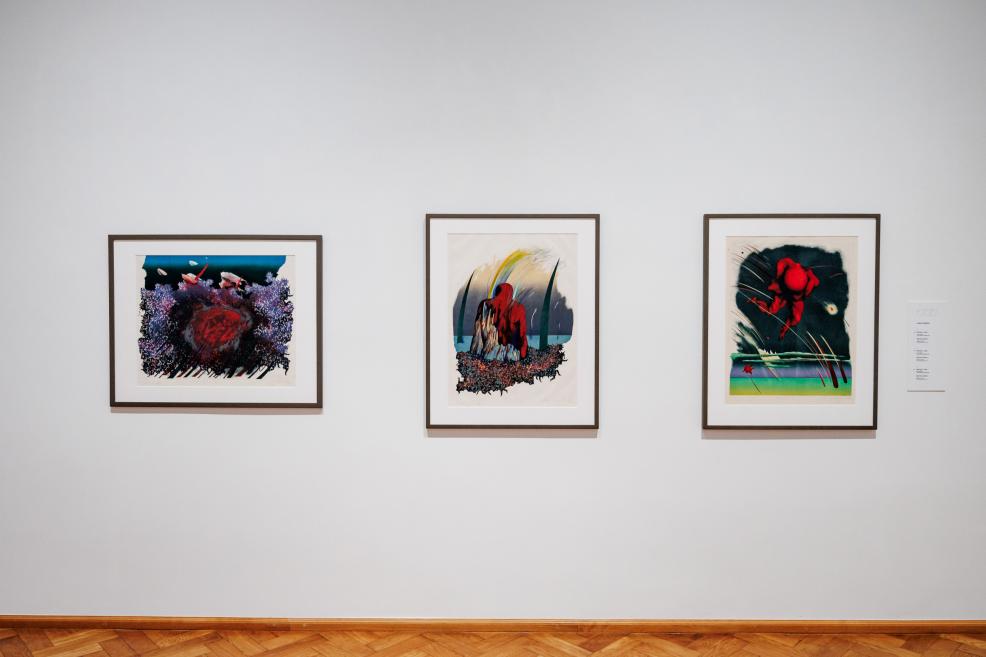 Three framed graphic artworks with expressive, vividly colored compositions hang on a white museum wall above a parquet floor.