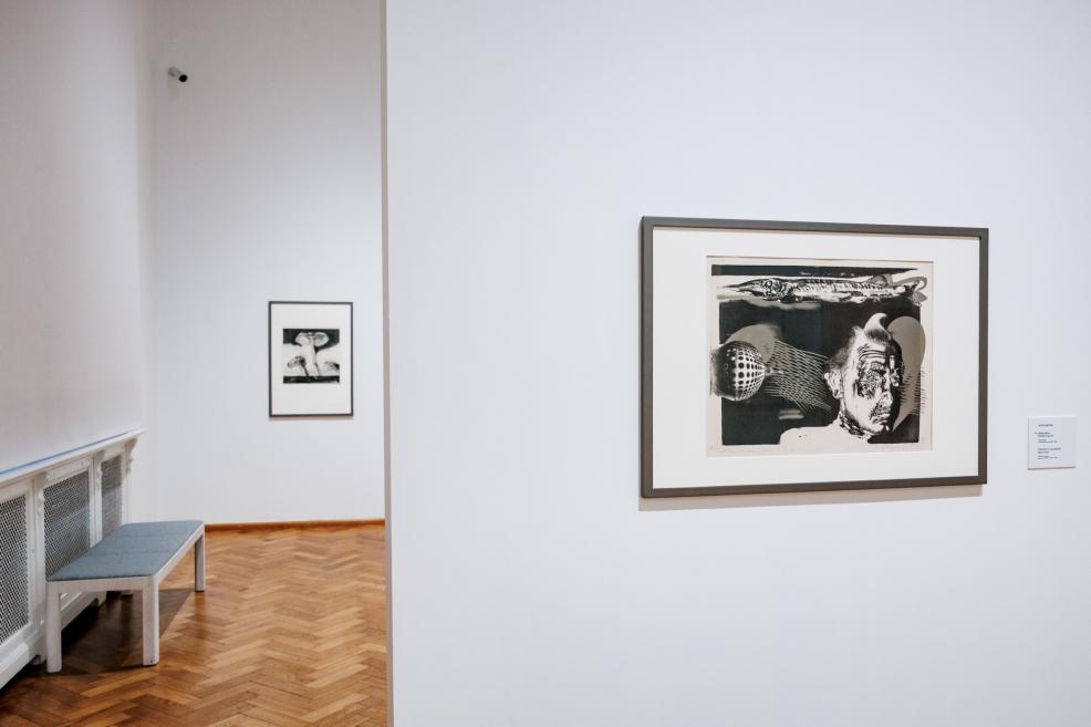In a museum gallery, two framed black-and-white graphic artworks are displayed on white walls, with a parquet floor and part of a bench visible.