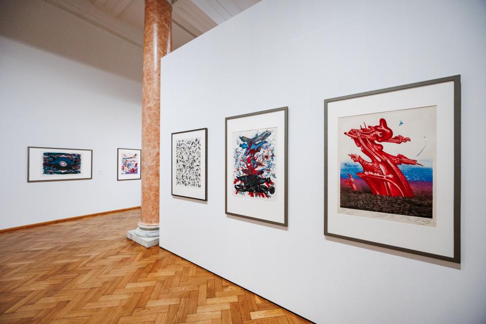 In a museum gallery, several framed colorful graphic artworks are displayed on a white partition wall, with a parquet floor and a marble column visible.