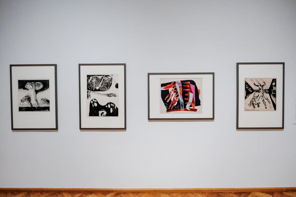 Uz baltas galerijas sienas vienā rindā izvietoti četri ierāmēti grafikas darbi ar atšķirīgām kompozīcijām.