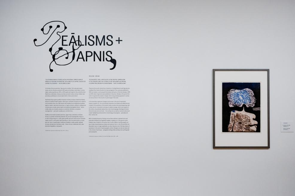 Museum gallery wall with the exhibition title “Reālisms + Sapnis” and explanatory text, alongside a framed graphic artwork.