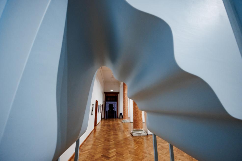 View of a museum gallery with parquet flooring and columns, photographed through the opening of a large white abstract sculpture.