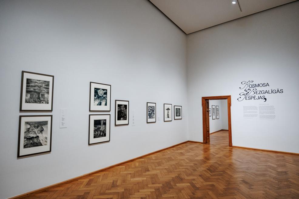 In a spacious museum gallery with parquet flooring, graphic artworks are displayed on the walls, and near the doorway the title “Infinite Possibilities of Space” marks the entrance to the next room.