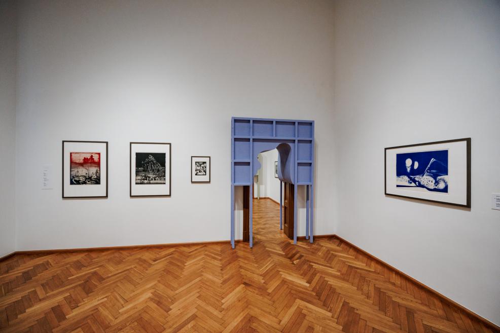 Muzeja zālē ar parketa grīdu pie sienas izvietoti trīs grafikas darbi, bet labajā pusē redzama zila telpiska konstrukcija ar atvērumu uz nākamo telpu.