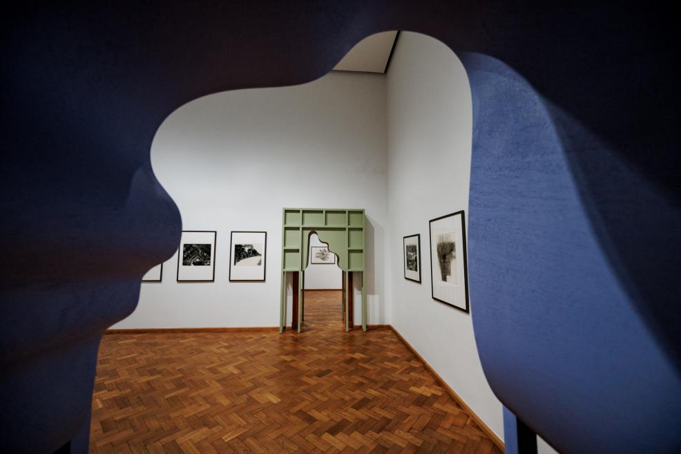 Exhibition hall with graphic artworks on white walls and a green architectural structure in the doorway, parquet floor.