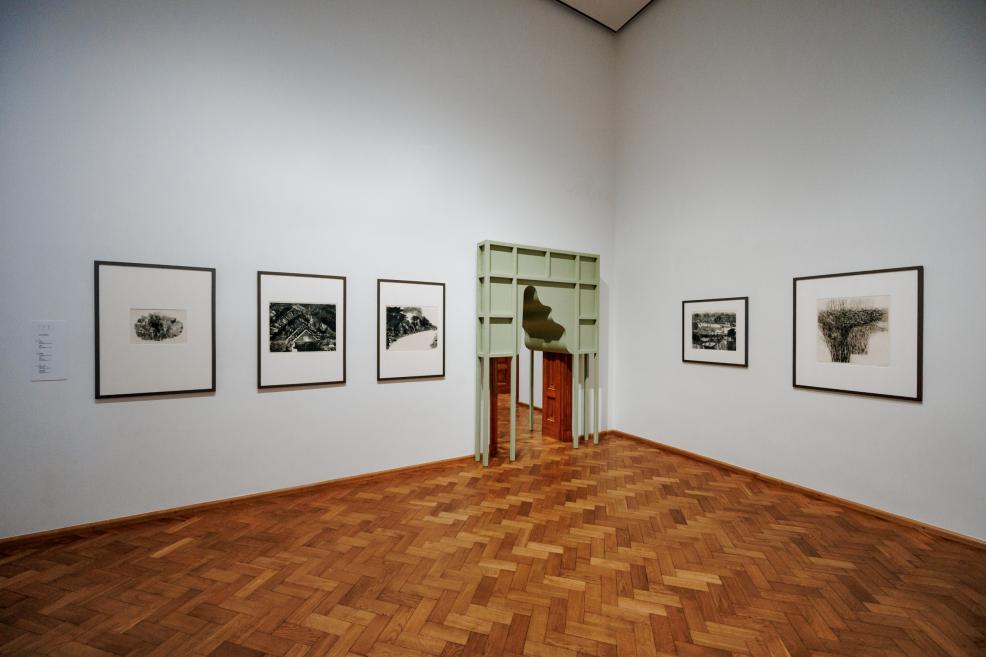 Exhibition hall with graphic artworks on white walls and a green architectural structure in the doorway, parquet floor.