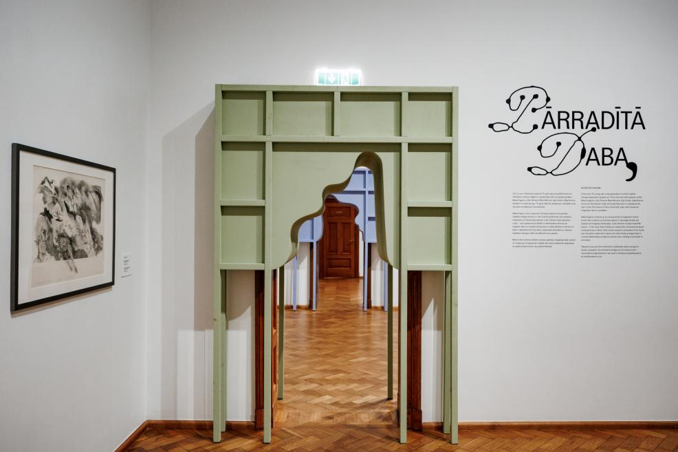 Zaļš dekoratīvs portāls ar viļņveida atvērumu galerijas telpā, blakus izstādes nosaukuma sienas tekstam un grafikām.