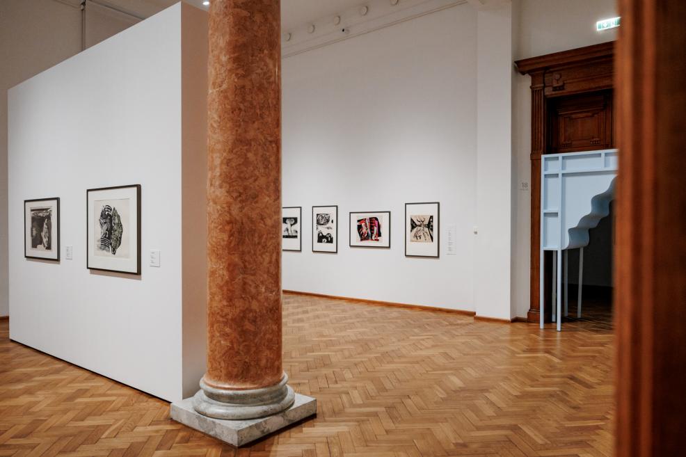 Exhibition hall with parquet flooring and a decorative marble column in the foreground, graphic artworks displayed on the walls and a light blue portal visible in the doorway.