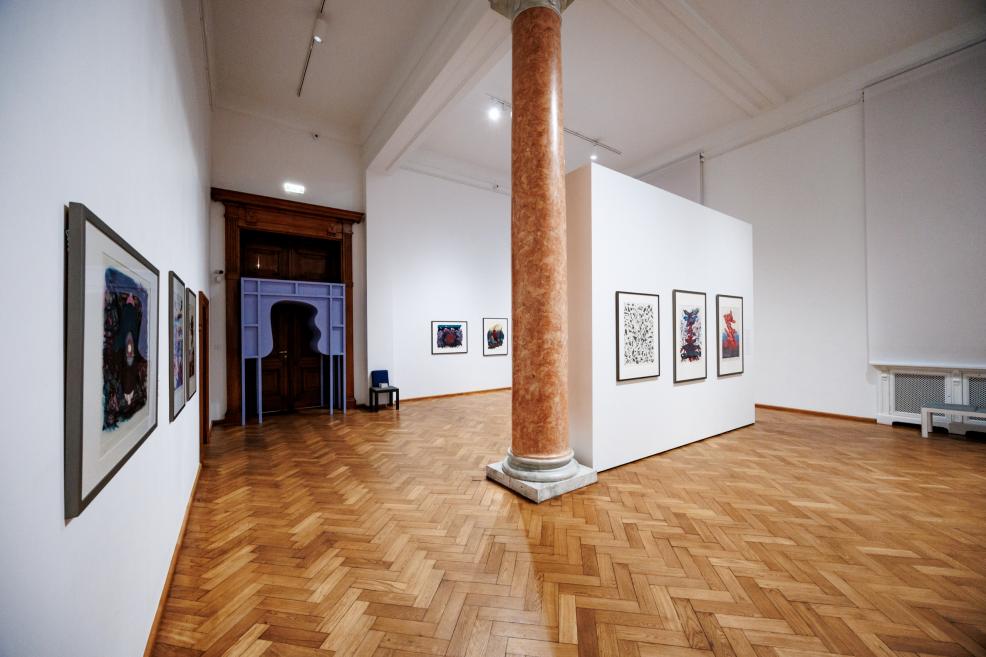 Spacious exhibition hall with parquet flooring and a central marble column, artworks displayed on the walls and a purple decorative portal visible in the doorway in the background.
