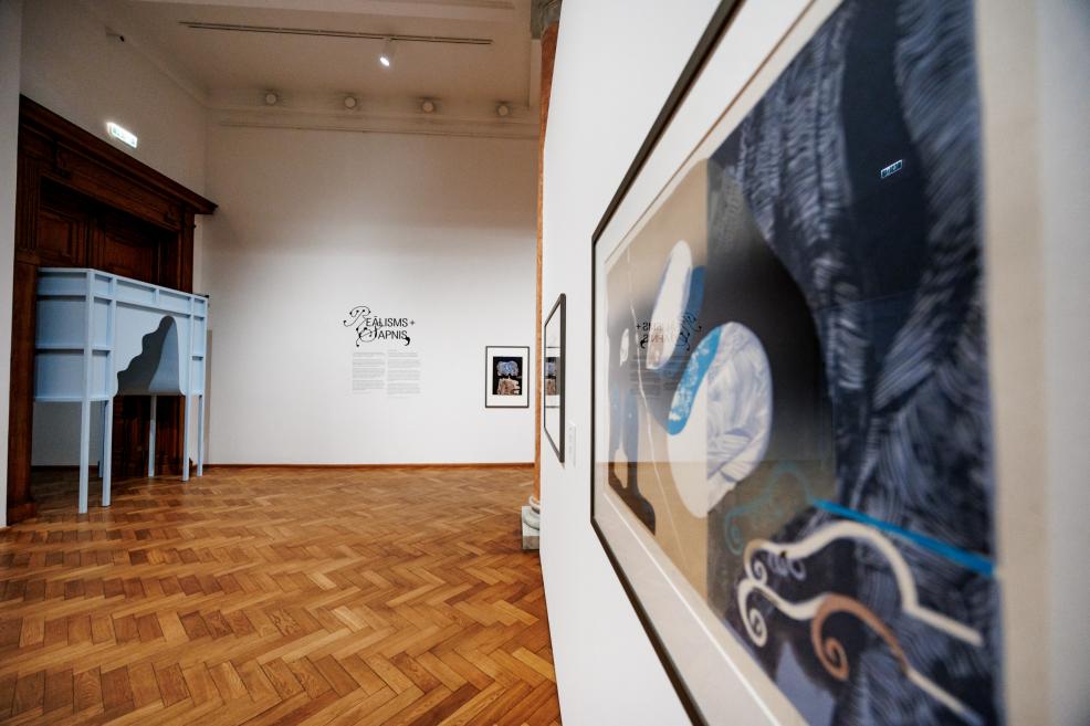 In the exhibition hall with parquet flooring, graphic artworks are displayed on the walls, with a framed piece in the foreground and an installation reading “Reālisms + Sapnis” visible in the distance.