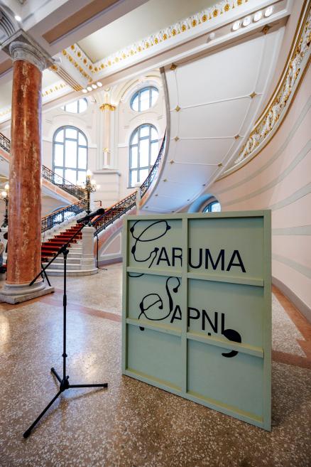 Latvijas Nacionālā mākslas muzeja centrālā vestibilā redzamas greznas kāpnes ar sarkanu paklāju, dekoratīvi griesti un kolonnas, priekšplānā novietots instalācijas elements ar uzrakstu “ĒĀLISMS + SAPNIS” un mikrofona statīvs.