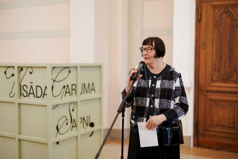 A woman with short dark hair and glasses stands at a microphone in a museum space holding a sheet of paper, with the exhibition title visible on a light green structure and wooden doors in the background.