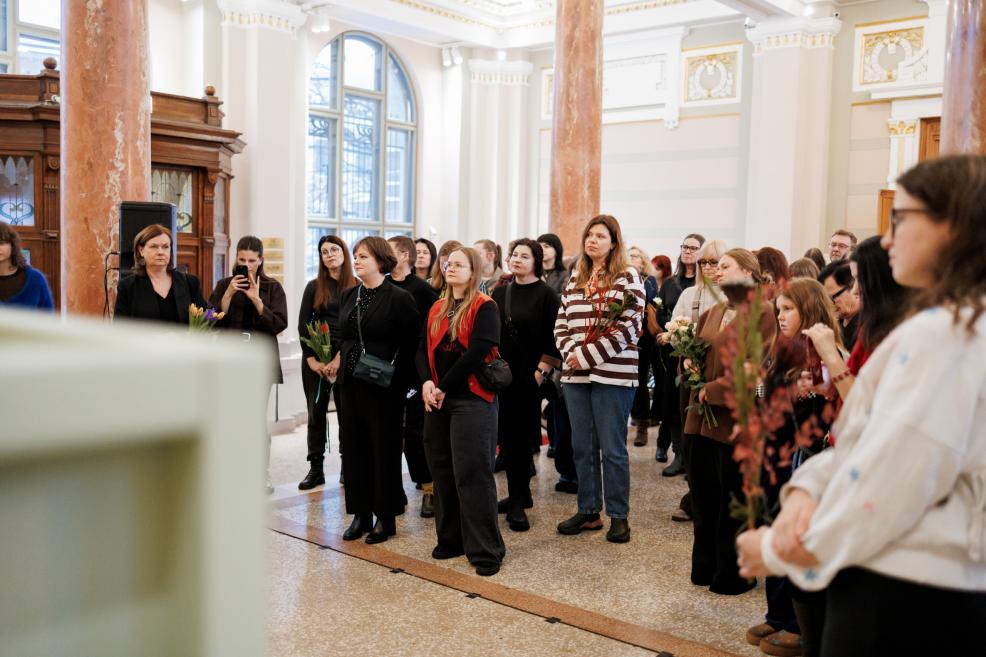 A group of people holding flowers listens to a speech in a museum lobby with tall windows, marble columns, and decorative ceilings.
