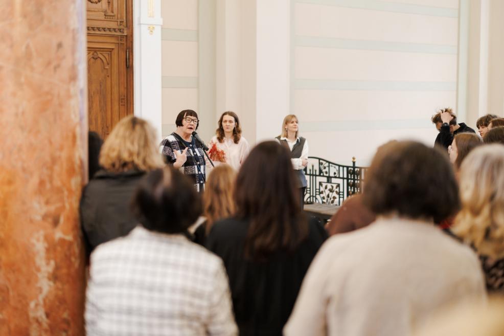 A woman with a microphone addresses the gathered audience in a museum lobby while listeners stand in the foreground with their backs to the camera.