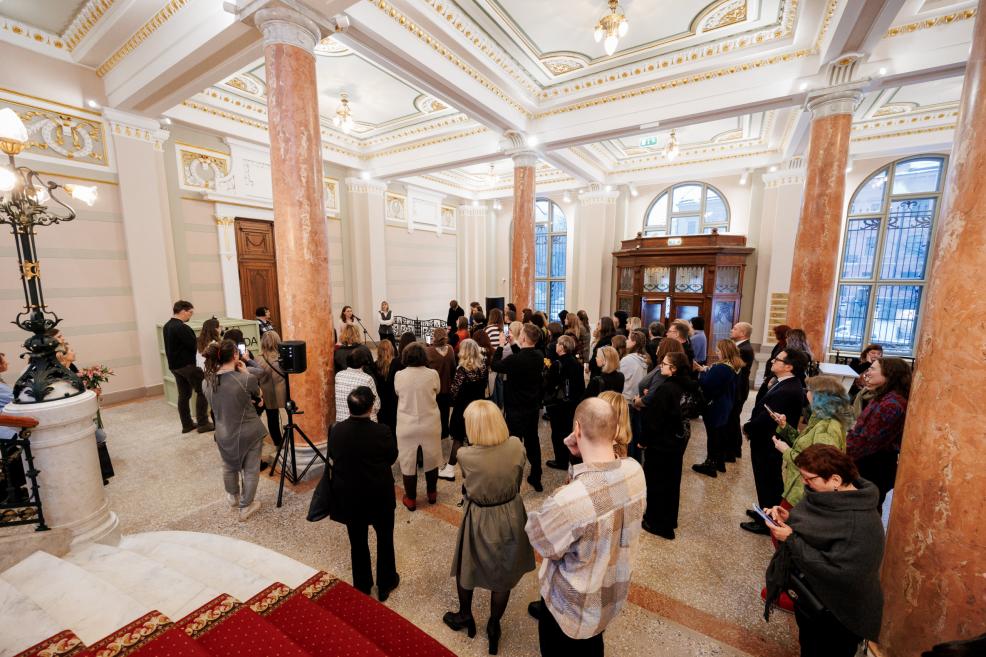 A wide view of a museum lobby with marble columns and ornate ceilings, where a crowd has gathered to listen to a speech.
