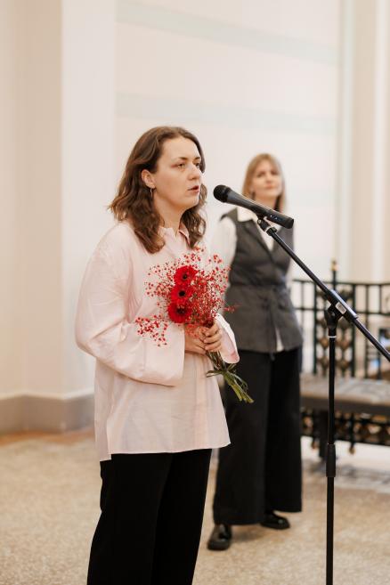A woman in a light blouse stands at a microphone holding a bouquet of red flowers, with another woman visible in the background of the museum lobby.