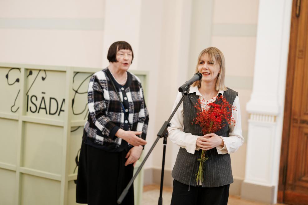 A woman in a gray vest stands at a microphone holding a bouquet of red flowers, with another woman visible behind her in the museum lobby.
