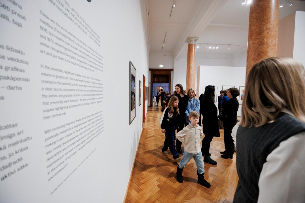 Visitors, including children, walk through a museum exhibition hall with paintings on the walls and informational text displayed on a white wall.