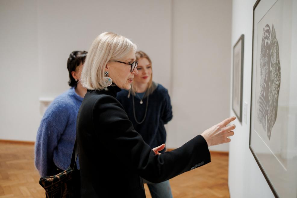A woman with glasses and light hair points at a graphic artwork on the museum wall while two other visitors observe it attentively.