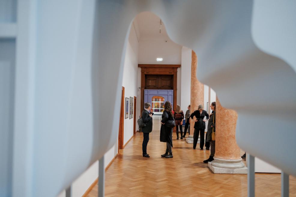 Through the opening of a sculpture, visitors can be seen conversing in a museum hall with parquet flooring and marble columns.