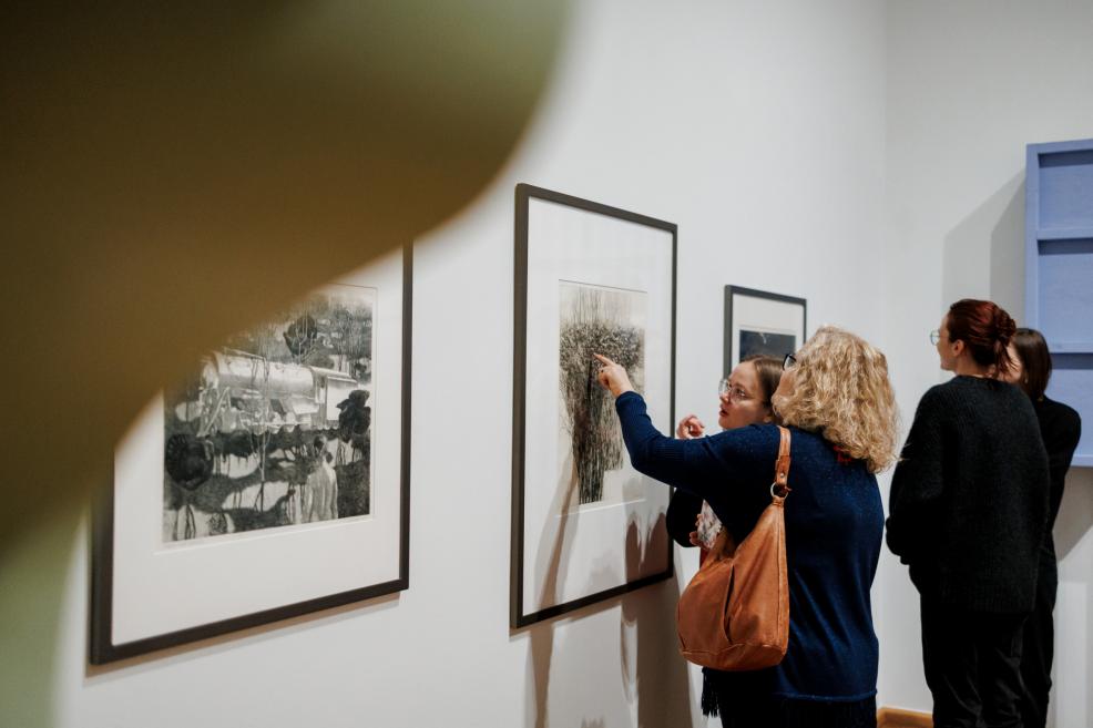 Visitors in a museum gallery observe graphic artworks, one of them pointing at a detail in the framed piece on the wall.