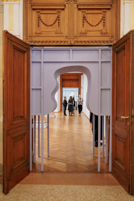 View through open wooden doors into a museum gallery featuring a light purple installation and visitors in the distance.