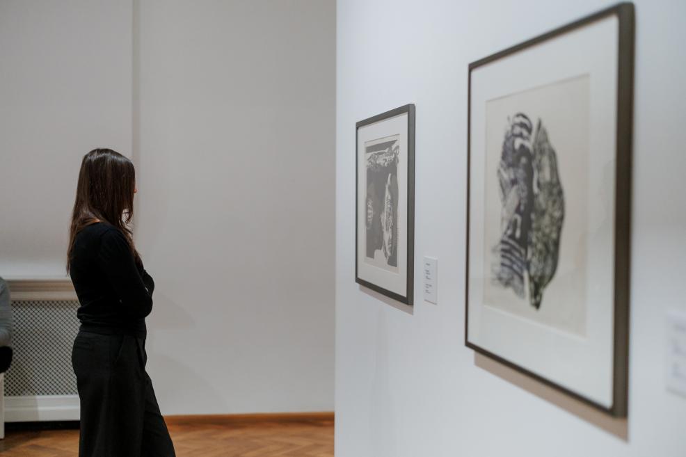 A woman stands by a white gallery wall observing two framed black-and-white prints.