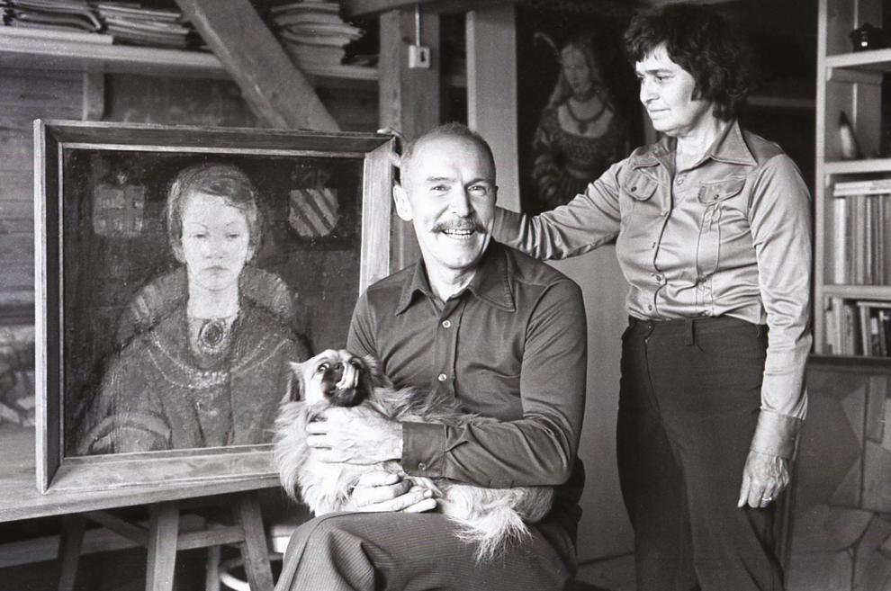 Black-and-white photograph of Ojārs Ābols seated in a studio with a dog, a woman and a portrait painting.
