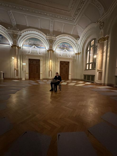 A spacious historic museum hall where a seated musician plays a hang drum, with exercise mats arranged around the room.