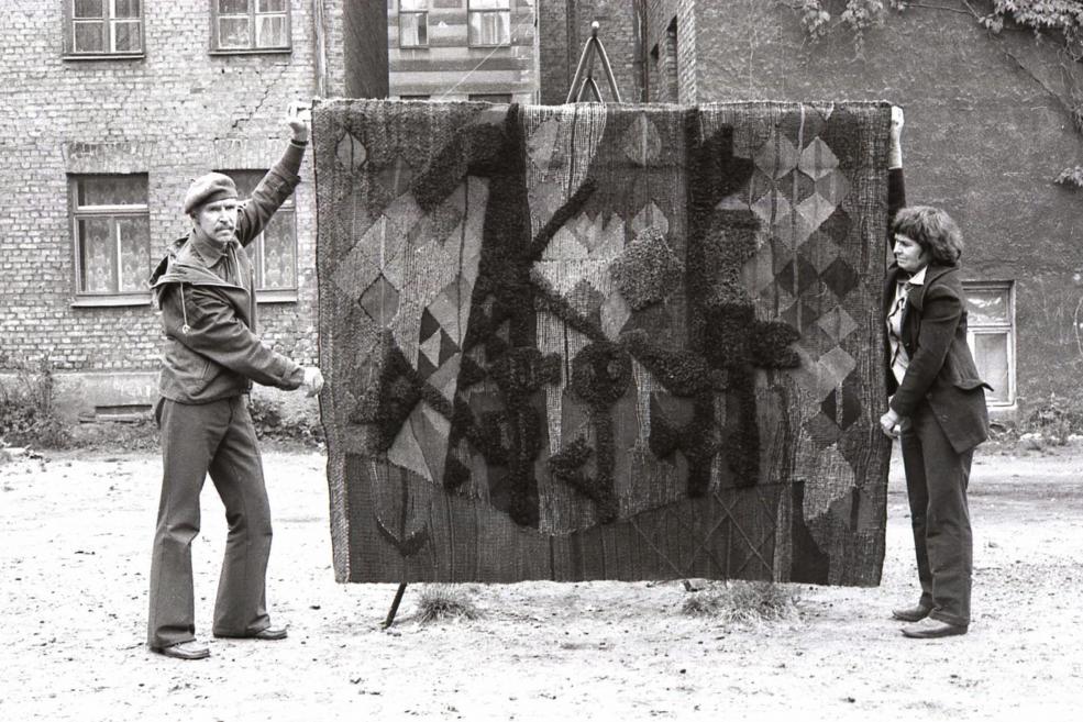 Black-and-white photograph of two people outdoors presenting a large textile artwork.