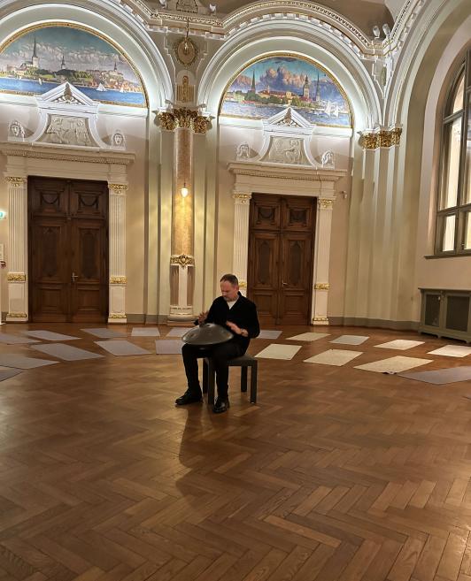 A musician sits on a chair playing a hang drum in an ornate museum hall.