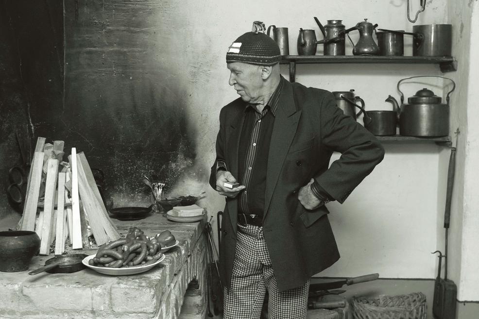 Black-and-white photograph of a man in a traditional interior beside a stove with kitchen utensils.