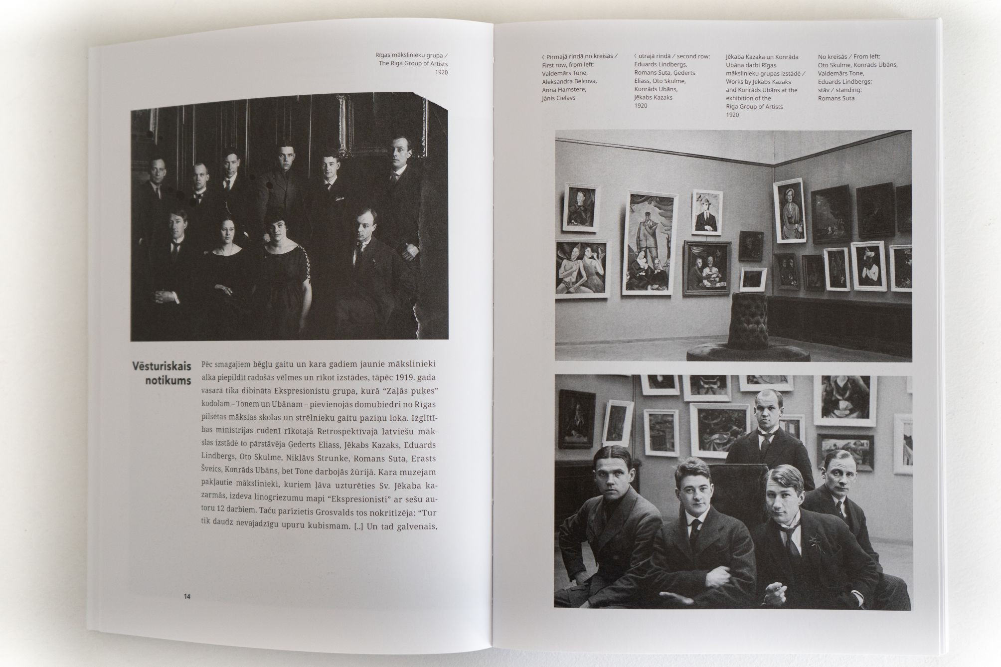 Atvērta grāmata ar melnbaltām fotogrāfijām – mākslinieku grupas portretu un izstādes skatiem ar gleznām pie sienām.