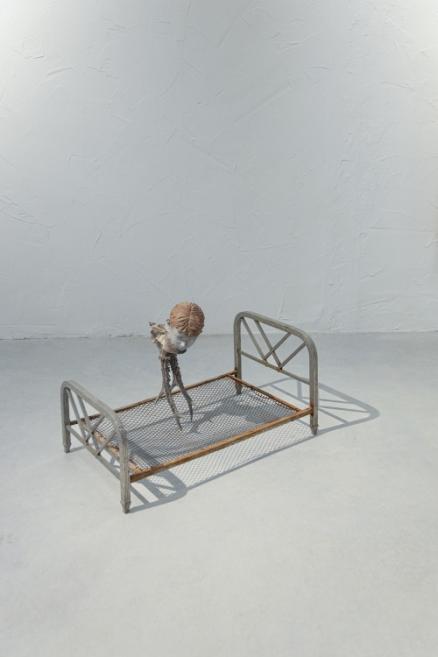 A metal bed frame stands on the floor with a small figurative sculpture placed on its mesh base against a white wall.