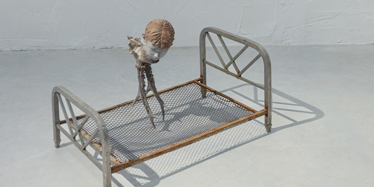 Sobre el suelo se encuentra una estructura met&aacute;lica de cama con una peque&ntilde;a escultura figurativa colocada sobre una base de rejilla junto a una pared blanca.