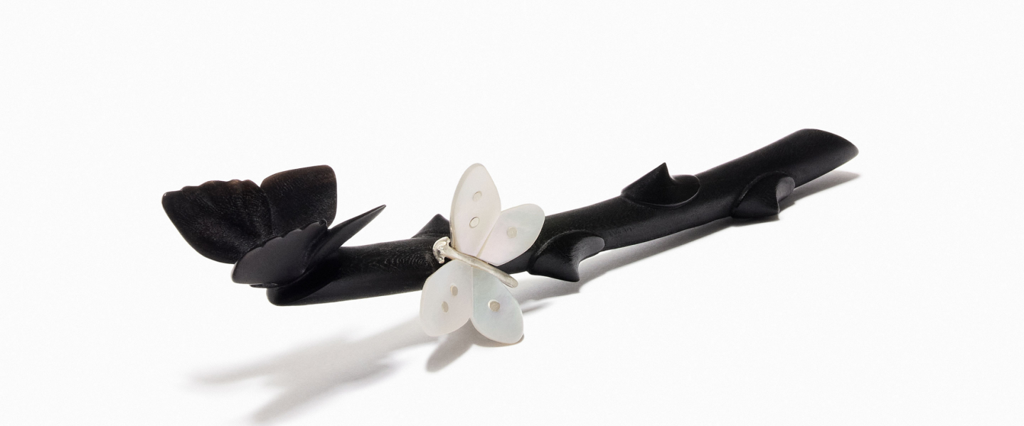 A jewellery piece shaped like a black branch with a stylised flower and a white butterfly, photographed against a light background.