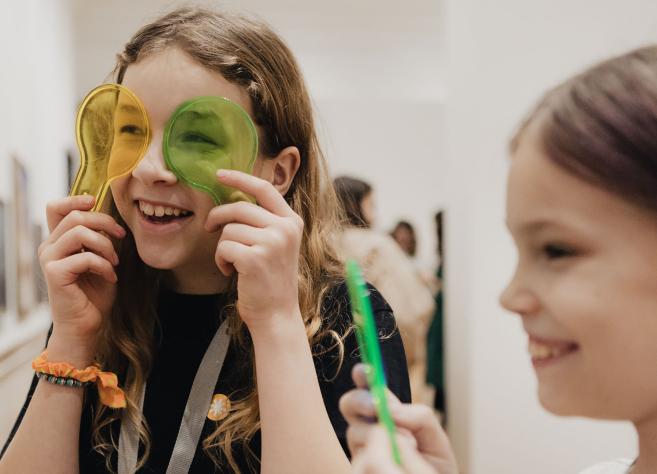 Divas meitenes muzeja zālē rotaļājas ar krāsainām plastmasas lēnēm, viena tur dzeltenu un zaļu priekšmetu pie acīm un smaida.