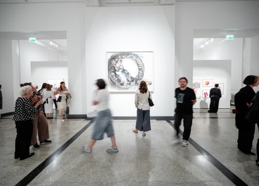 Muzeja izstāžu zāle ar baltām sienām, kur apmeklētāji skatās uz lielu sienas darbu centrā un vairākiem ierāmētiem darbiem otrā zālē.