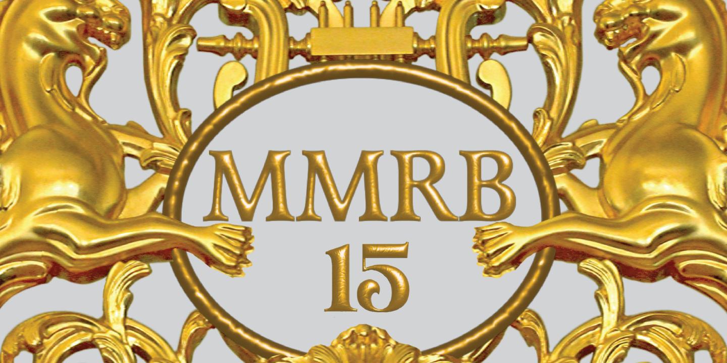Zeltīts dekoratīvs ornaments ar diviem spārnotiem grifiem, vīnogu ķekariem un centrālu medaljonu ar uzrakstu “MMRB 15”.