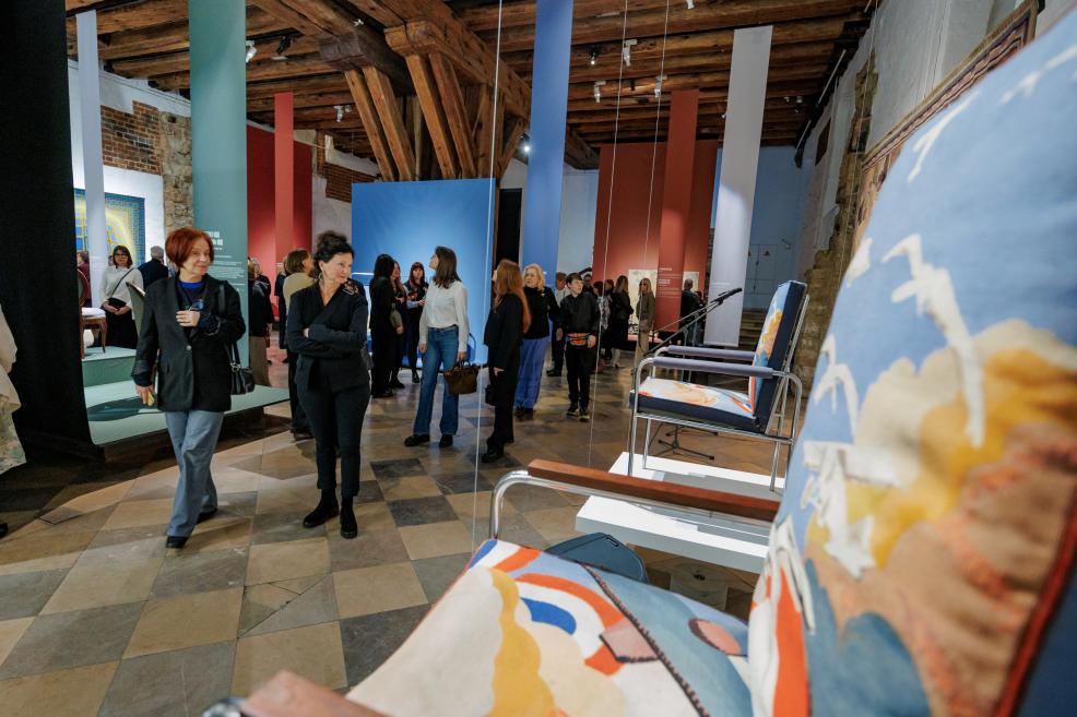 Muzeja izstāžu zālē apmeklētāji pārvietojas starp eksponātiem, tostarp dekoratīviem krēsliem un mākslas darbiem.