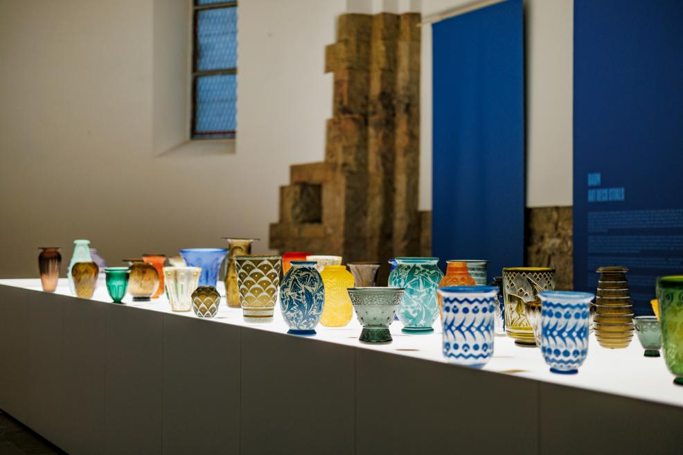 Uz izgaismota galda muzeja telpā izstādīti dažādu formu un krāsu stikla trauki ar dekoratīviem rakstiem.